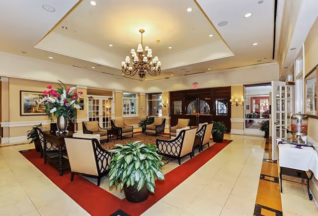 A spacious and elegant common area with beige walls and tiled floors, featuring a red carpeted seating area with multiple cushioned chairs arranged around a central rug. There are large potted plants, a chandelier hanging from the ceiling, framed artwork on the walls, and double wooden doors in the background. A table with a water dispenser and cups is visible on the right side.