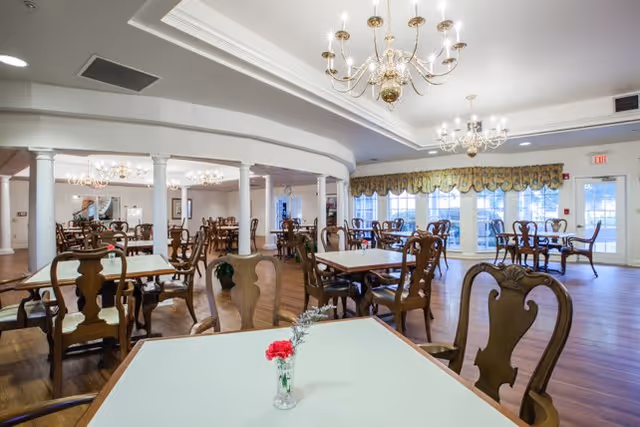 Spacious dining room with multiple wooden tables and chairs arranged neatly. The room features large windows with decorative valances, elegant chandeliers hanging from the ceiling, and a polished wooden floor. A small vase with a red flower is placed on the closest table.
