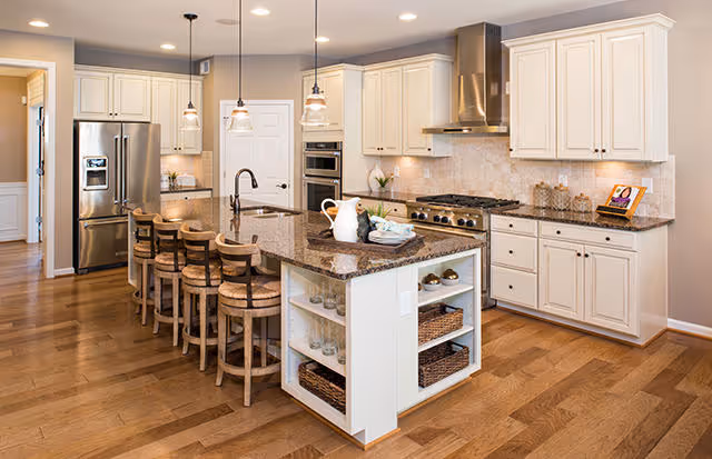 A spacious modern kitchen featuring a large island with a granite countertop and four wooden bar stools. The kitchen has white cabinetry, stainless steel appliances including a refrigerator and a stove with a range hood, pendant lighting above the island, and hardwood flooring.
