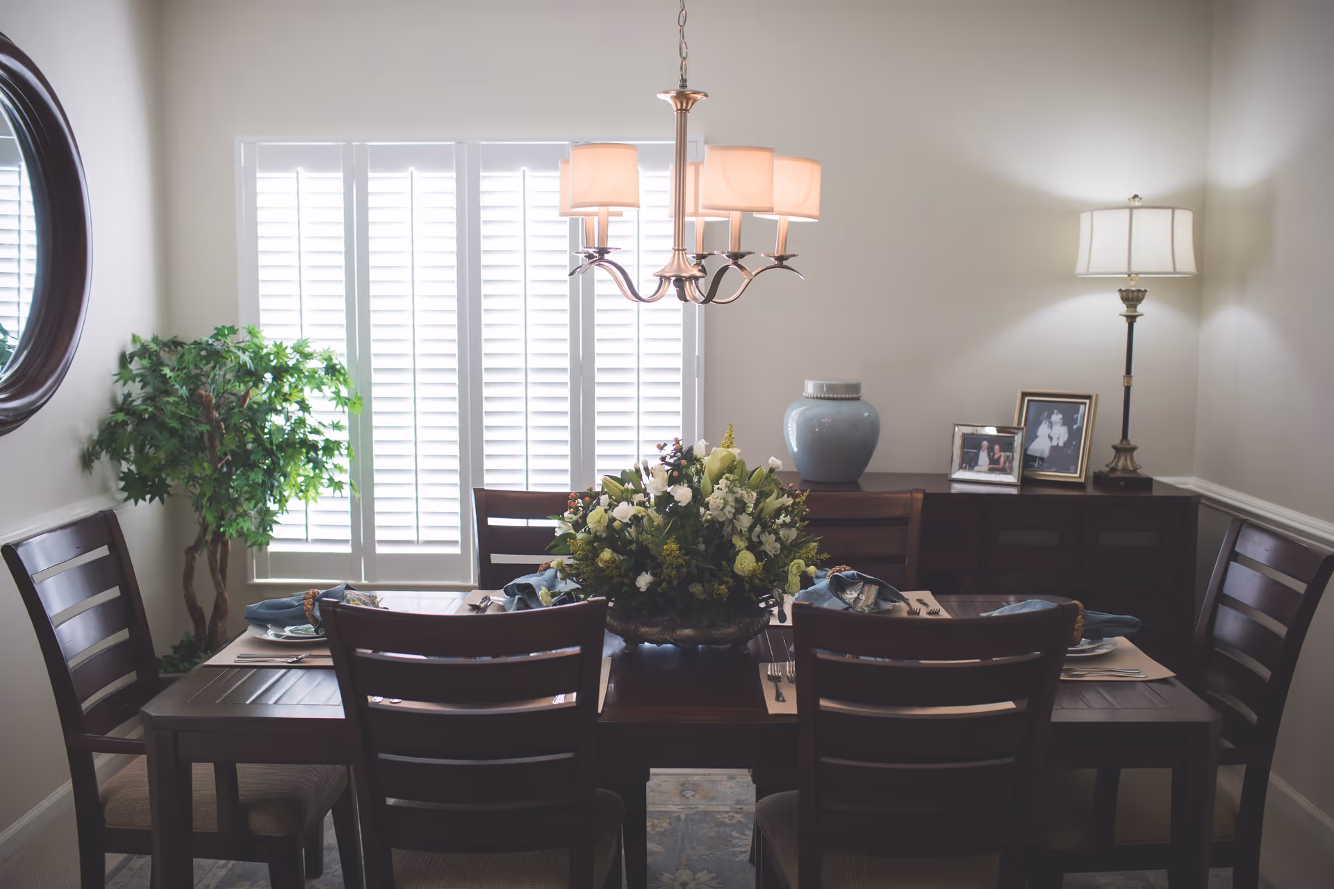 Dining room with a wooden table set for six, a floral centerpiece, chandelier, sideboard with a lamp and framed photos, and shuttered windows.