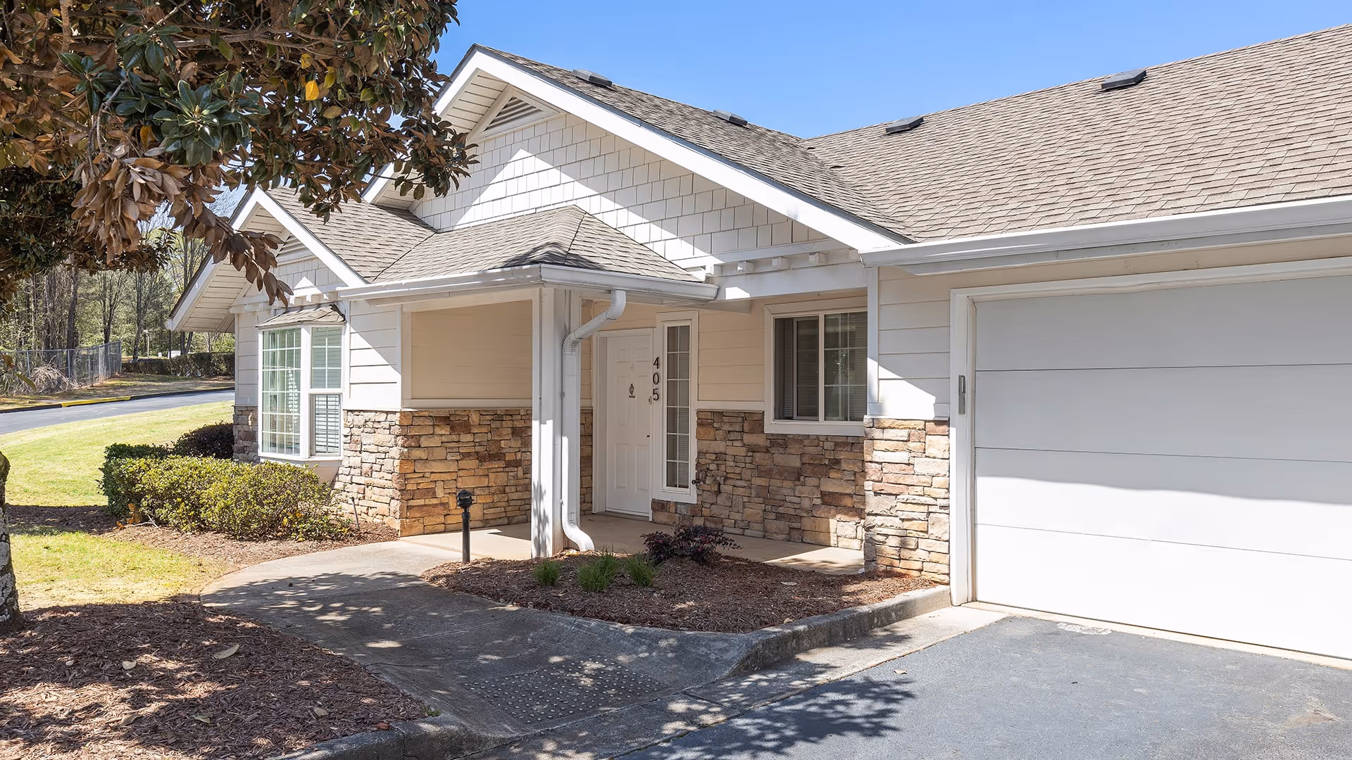Exterior view of a single-story residential building with white siding and stone accents. The entrance features a covered porch with the number 405 on the door. There is a garage door to the right and landscaping with bushes and a tree casting shadows on the driveway and walkway.