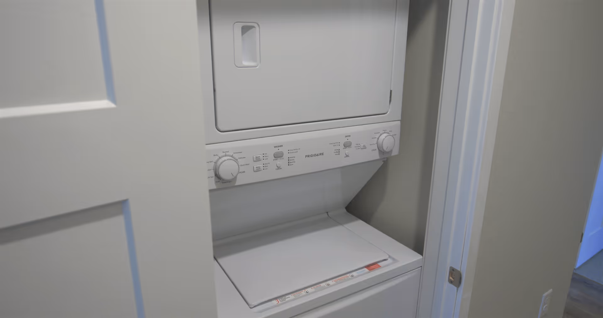 A white stacked washer and dryer unit partially enclosed in a small laundry closet with a white door slightly open.