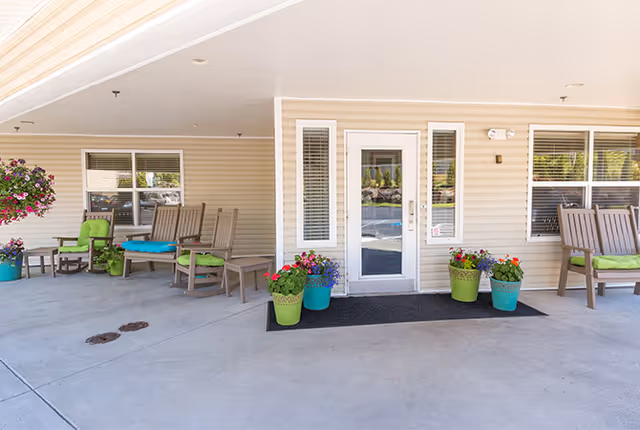 Covered front entrance with a glass door, wooden chairs and colorful potted flowers on a concrete porch.