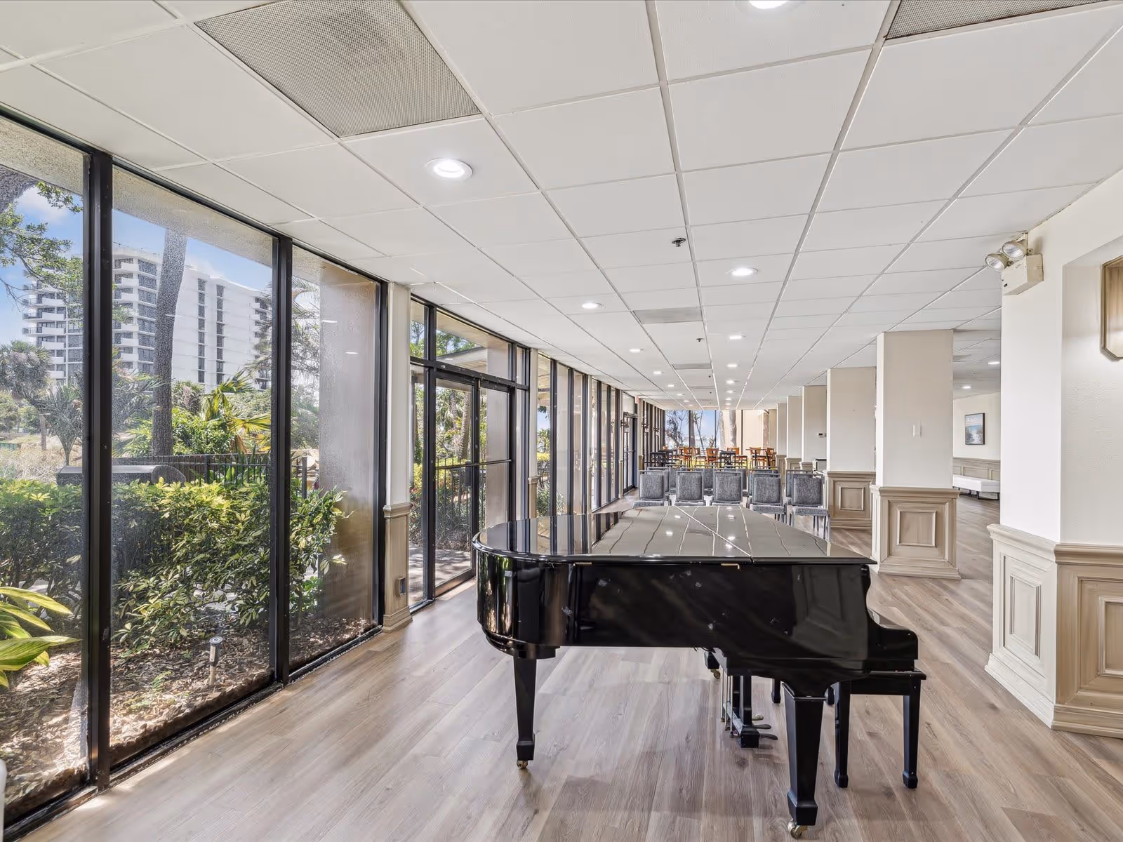 A spacious interior room with large floor-to-ceiling windows on the left side showing greenery and buildings outside. In the center of the room is a black grand piano. Further back, there are rows of chairs and tables arranged in a dining or meeting area. The room has light wood flooring and a white drop ceiling with recessed lighting.