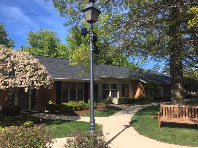 Outdoor view of a senior living facility with a paved walkway, a wooden bench, a lamppost, blooming trees, and a brick building with windows and a black roof under a clear blue sky.