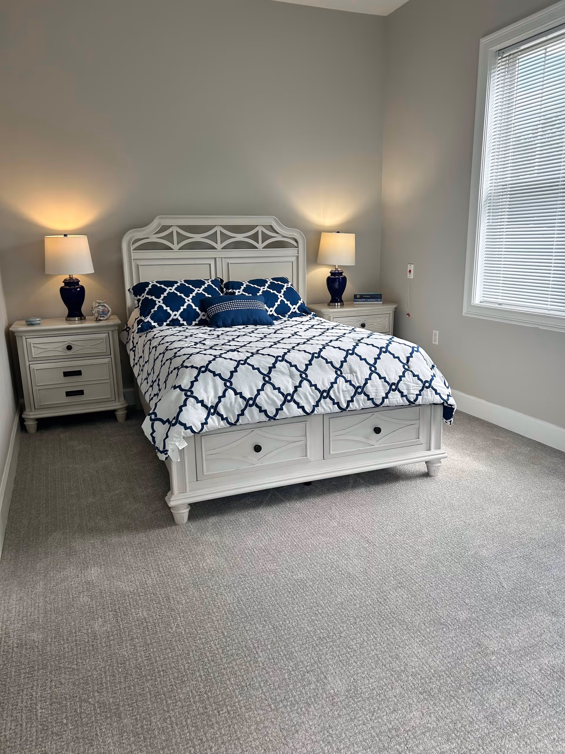 A neatly made bed with a white and navy blue patterned comforter and matching pillows in a bedroom. The bed has a white wooden headboard and footboard with storage drawers. There are two white nightstands on either side of the bed, each with a blue lamp. A window with white blinds is on the right wall, and the room has light gray walls and carpeted flooring.