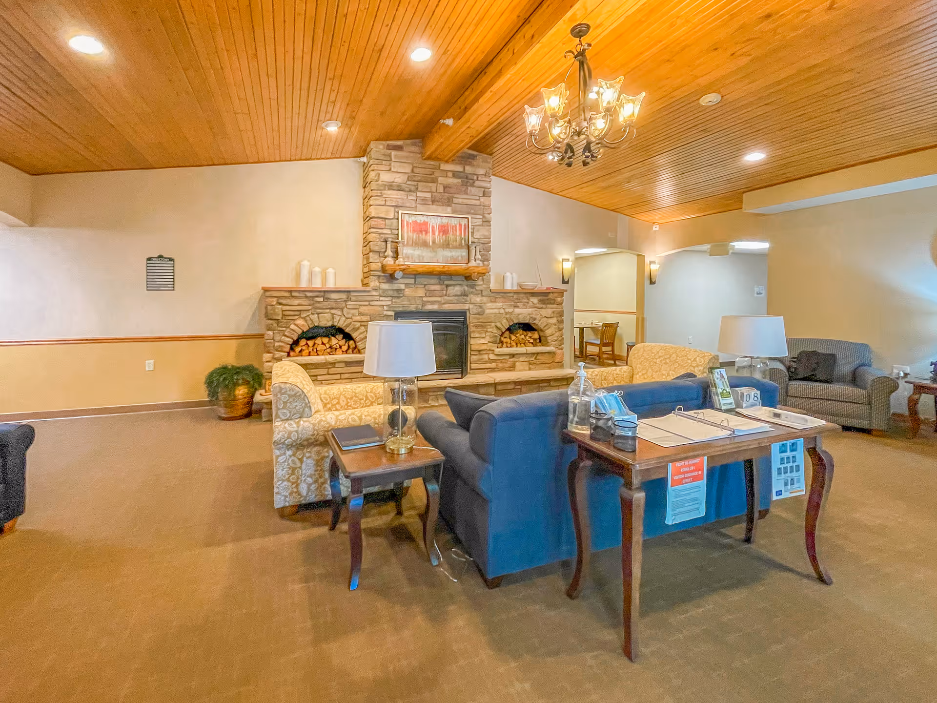 Communal lounge with sofas and chairs arranged around a stone fireplace beneath a wood-paneled ceiling.