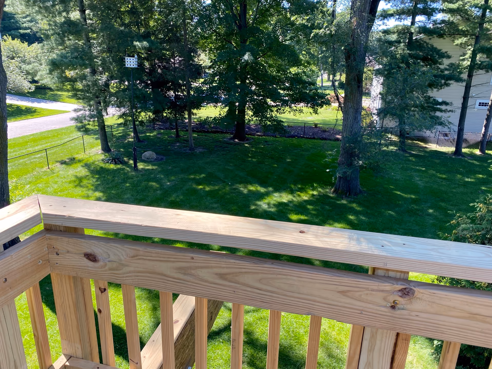 View from a wooden balcony railing overlooking a grassy backyard with trees and a birdhouse mounted on a pole.