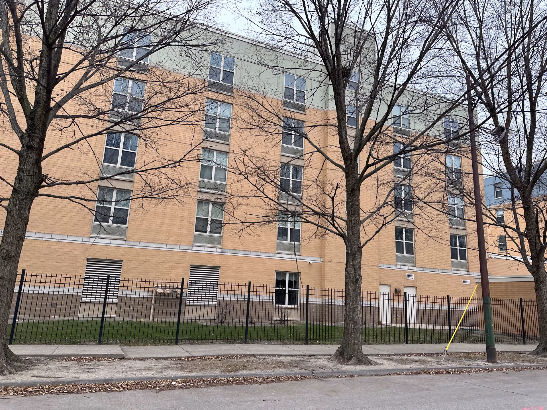 Brick multi-story building facade with rows of windows, a black metal fence, and leafless trees along the sidewalk.