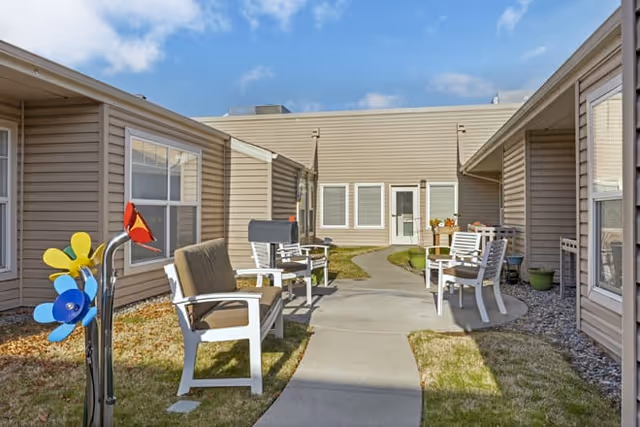 Sunny courtyard with a curved concrete path lined by white outdoor chairs between beige single-story buildings.