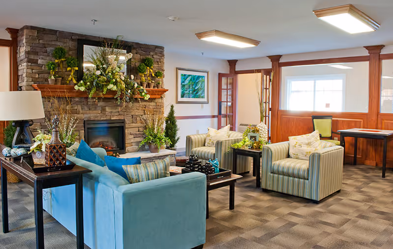 A cozy living room area in a senior living facility with a stone fireplace decorated with plants and flowers. The room features a light blue sofa with striped and solid pillows, two striped armchairs, a wooden coffee table, and side tables with decorative items. The walls have wood paneling and framed artwork, and the room is well-lit with ceiling lights and natural light from a window.
