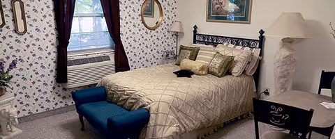 A cozy bedroom with a neatly made bed featuring multiple pillows and a beige quilt. The room has floral wallpaper, a window with dark curtains, an air conditioning unit below the window, a blue upholstered bench at the foot of the bed, a round mirror on the wall, a floor lamp, a framed picture above the bed, and a small table with a lamp and chair.