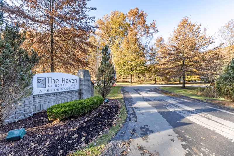 Entrance sign for The Haven at North Hills Senior Residence surrounded by landscaped bushes and trees with autumn foliage, next to a paved driveway.