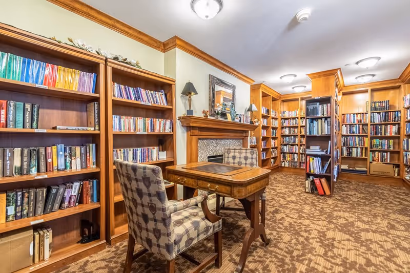 A cozy library room with wooden bookshelves filled with books lining the walls. In the center, there is a wooden table with a chessboard on top and two patterned upholstered chairs. The room features a fireplace with a decorative mirror and lamps on the mantel, and the floor is carpeted with a patterned design.