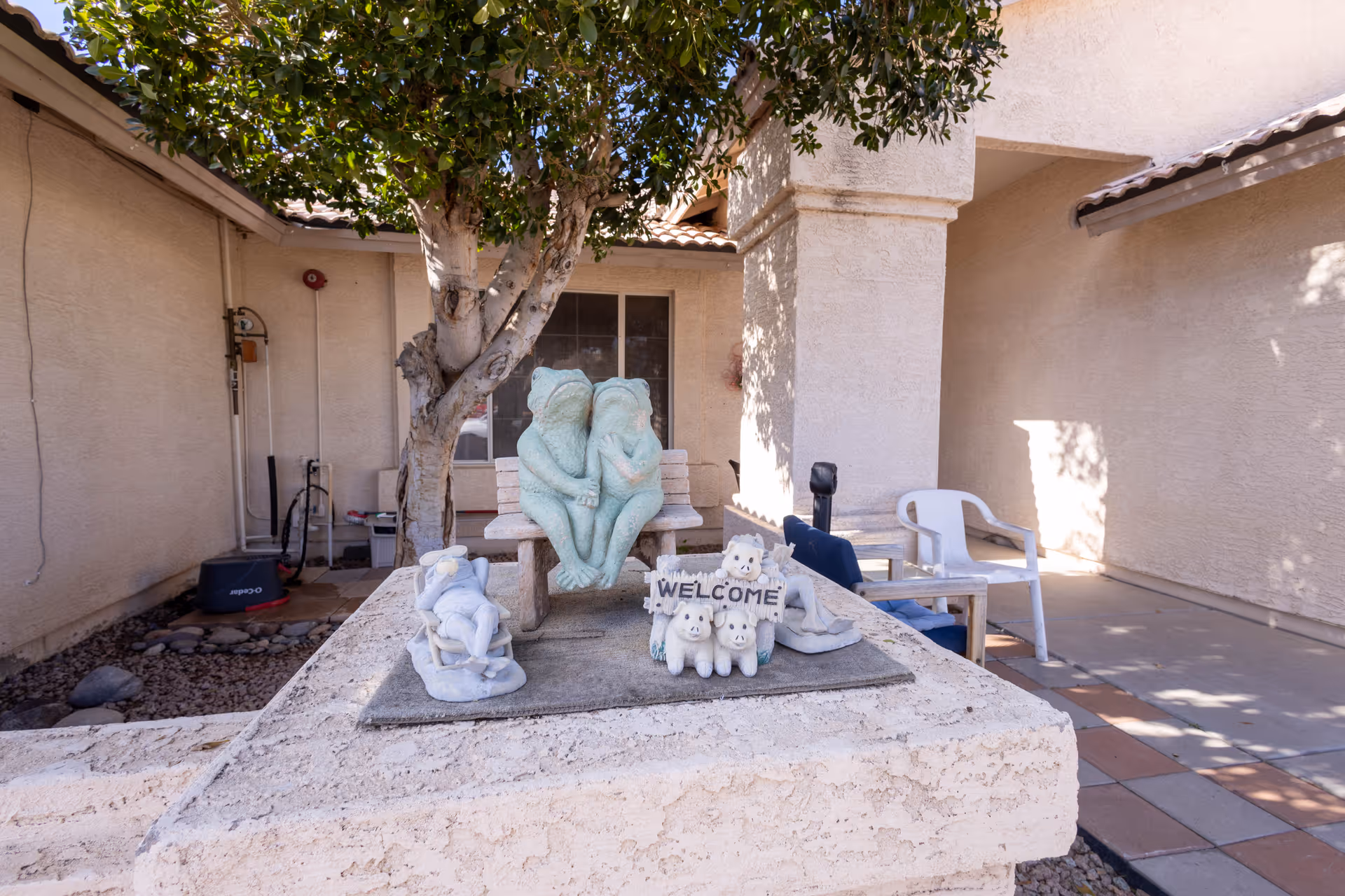 Outdoor courtyard area with a stone table displaying decorative statues including two frogs sitting on a bench, a 'WELCOME' sign with small animal figures, and other small figurines. There is a tree behind the table, a white plastic chair, and beige stucco walls surrounding the space.