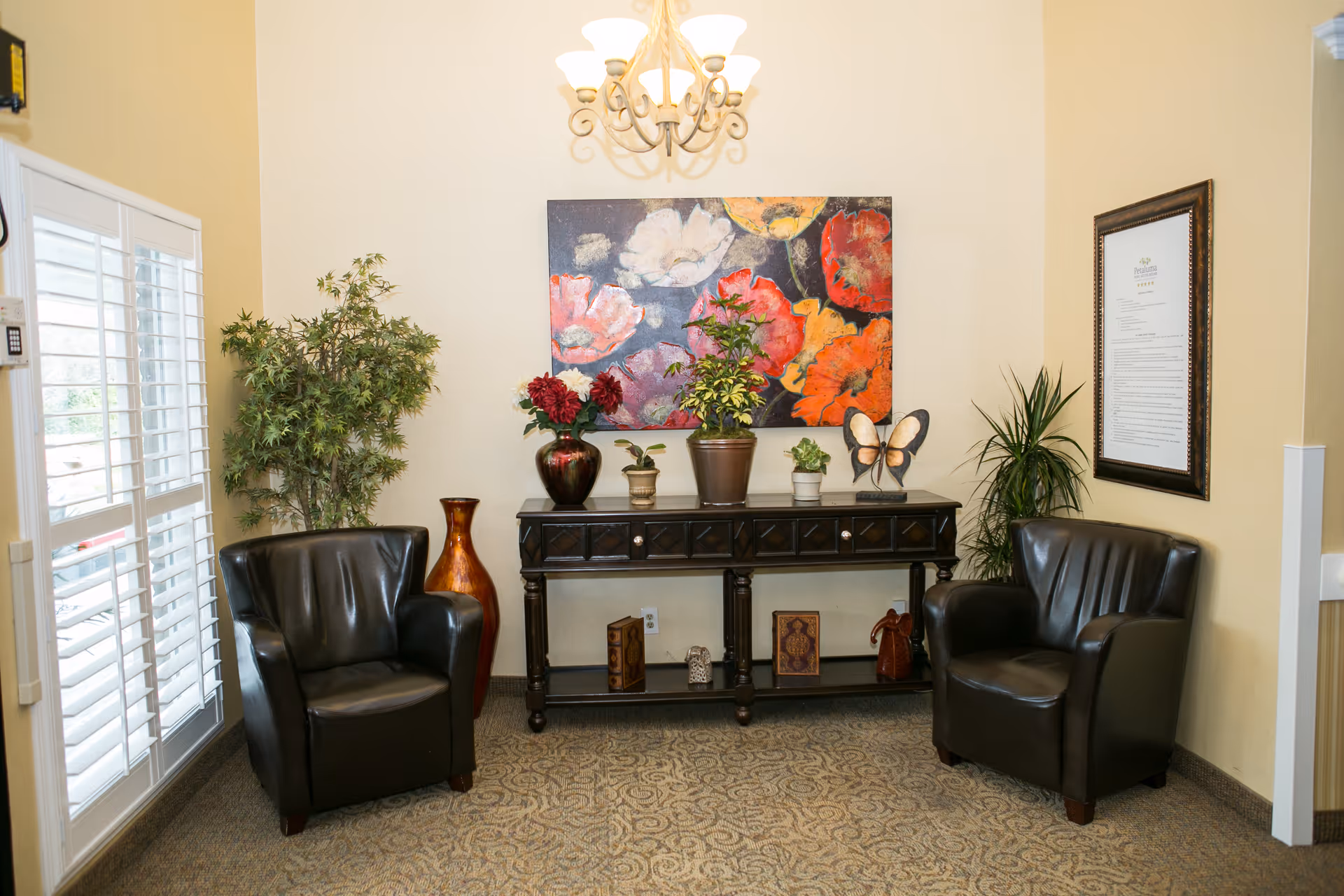 Two black leather armchairs flank a decorative console table topped with plants and a colorful floral painting on the wall in a cozy sitting area.