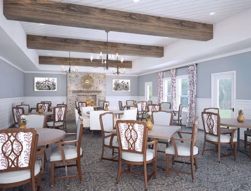 A bright dining room with multiple wooden tables and chairs featuring patterned upholstery. The room has a carpeted floor with a floral design, light gray walls with white wainscoting, and large windows with floral curtains letting in natural light. There are two chandeliers hanging from the ceiling with exposed wooden beams. A brick fireplace with a decorative mirror above it is centered on the far wall, flanked by two framed artworks.