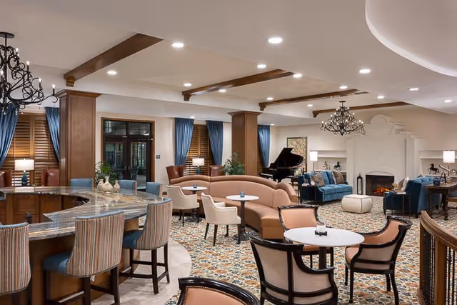 A spacious and elegantly furnished senior living common area featuring a curved bar with high chairs, multiple seating arrangements including armchairs and sofas, a grand piano, and a fireplace. The room has wooden beams on the ceiling, blue curtains, and a patterned carpet.