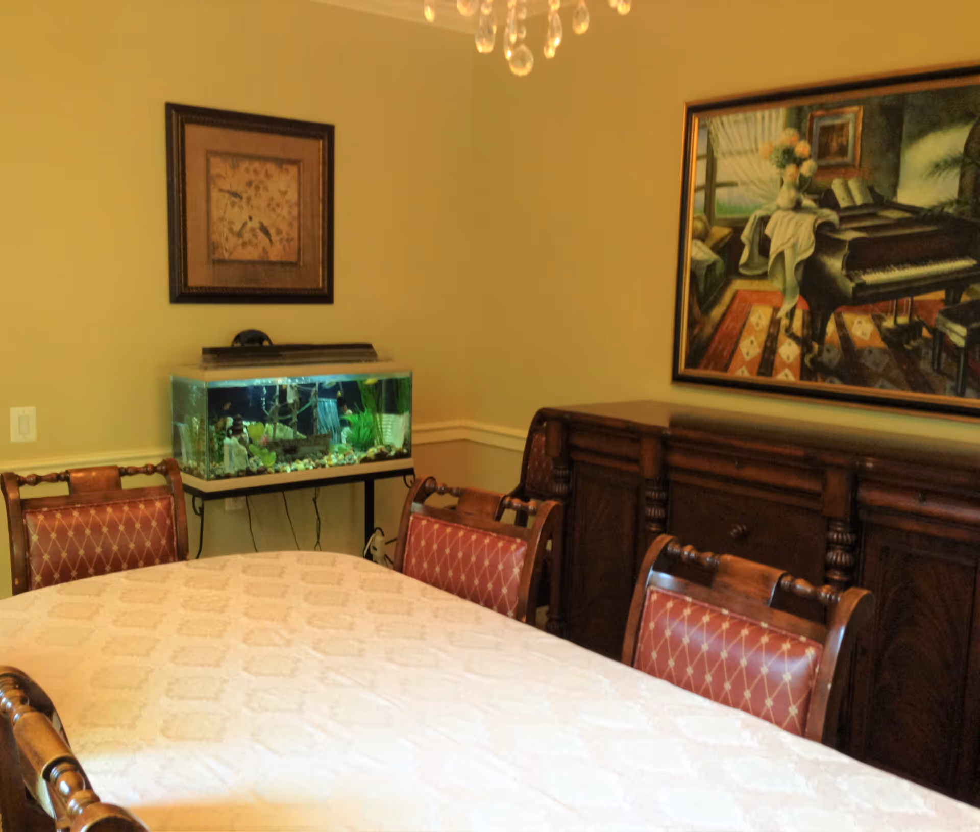 Dining room with a table covered by a white tablecloth and four wooden chairs with red cushioned seats. There is a fish tank on a stand against the wall, a wooden sideboard, and two framed paintings hanging on the walls. A chandelier with hanging crystals is partially visible above the table.