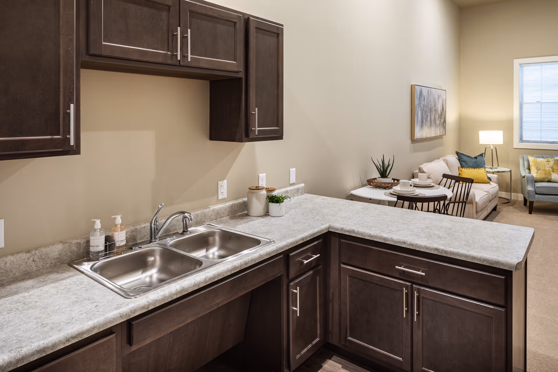 Interior view of a living space in Boyson Heights Assisted Living featuring a kitchen area with dark wood cabinets, a double stainless steel sink, and a light-colored countertop. In the background, there is a small round dining table set with dishes, a beige sofa with colorful pillows, a side table with a lamp, and a window with blinds.