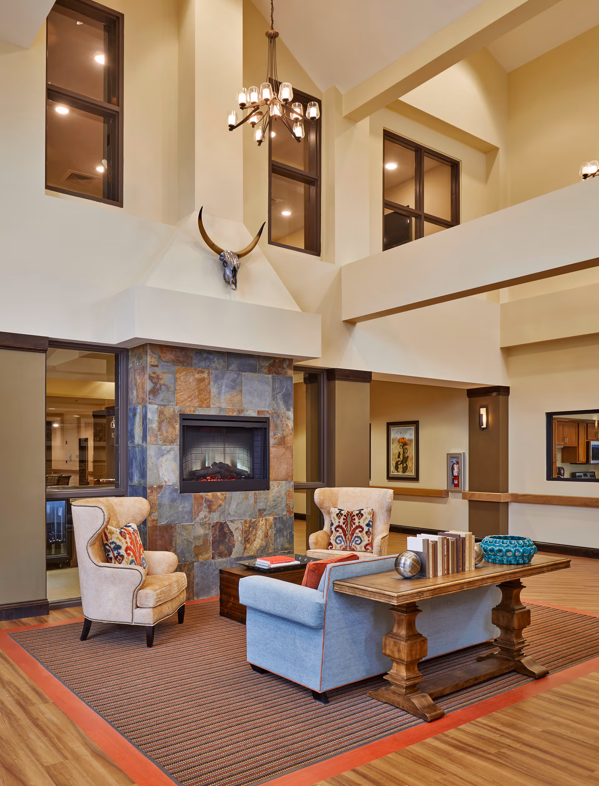 High‑ceilinged lounge area with armchairs and a sofa arranged around a stone fireplace, a wooden table, chandelier, and decorative skull on the wall.
