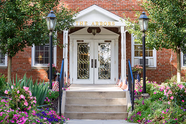 Entrance to The Arbors building with white double doors featuring decorative glass panels, flanked by two black lamp posts and surrounded by green trees and colorful flowers. The building is made of red brick and has a small set of concrete steps leading up to the doors.