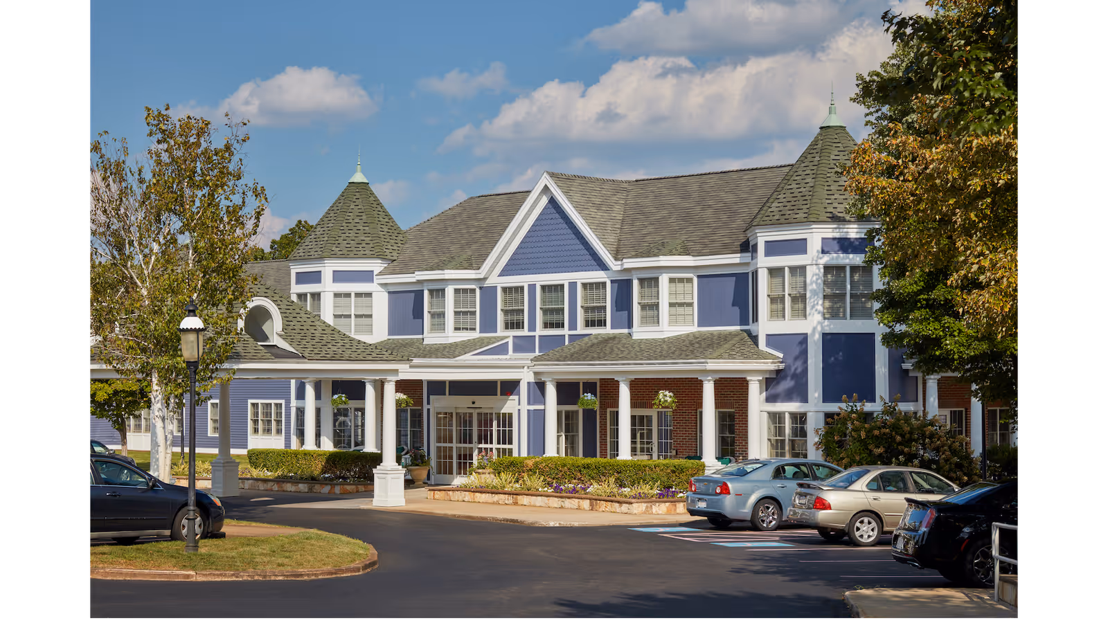 Front entrance of a large blue-and-white assisted living building with a covered porte-cochere, landscaped beds, and parked cars in the driveway.