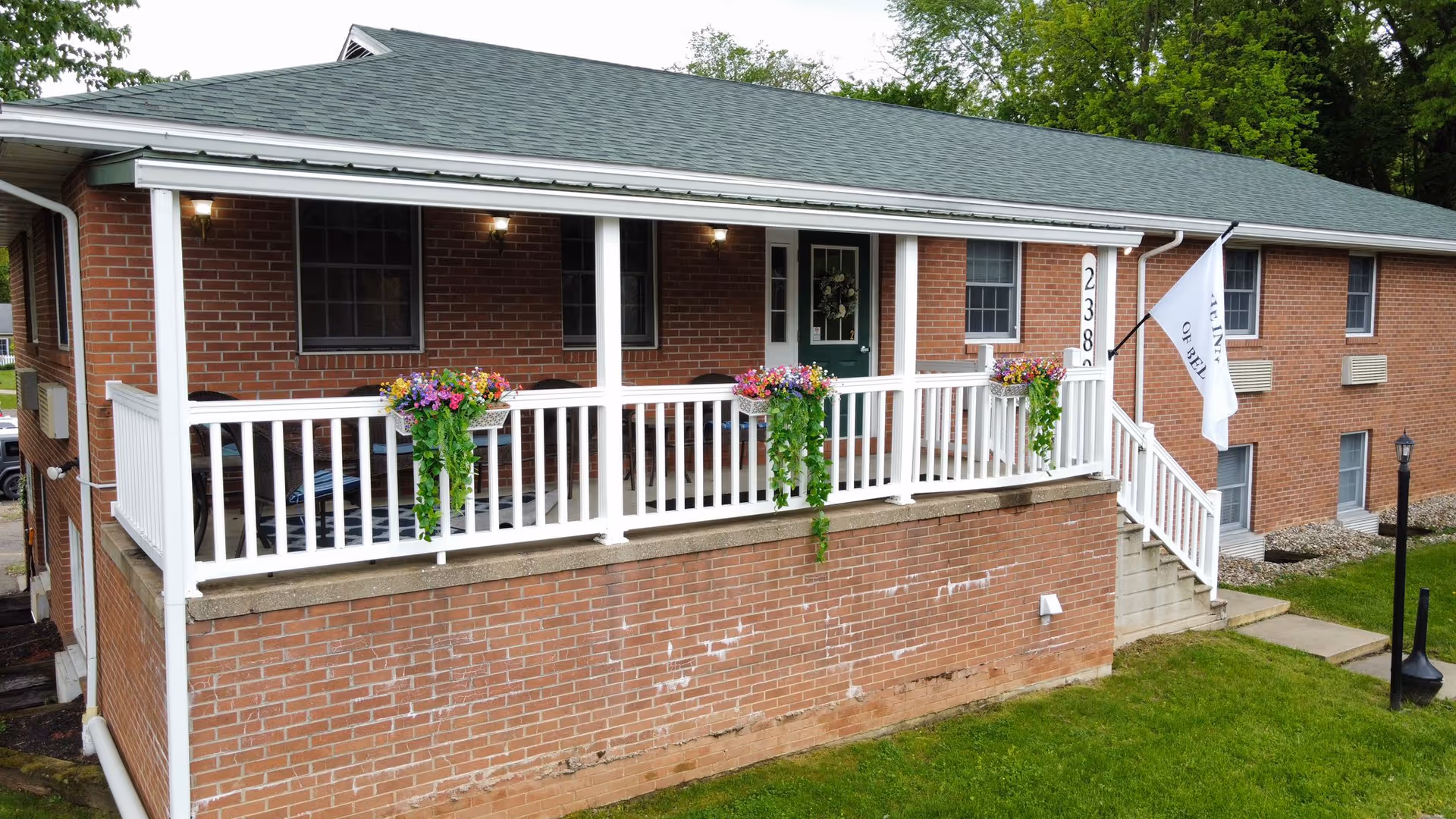 Front exterior of a brick senior living building with a covered porch, white railing, flower boxes, and an entrance door.
