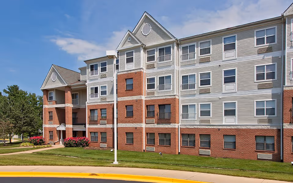 Exterior view of a multi-story senior living facility building with a combination of red brick and light-colored siding, surrounded by green grass and a few trees under a clear blue sky.