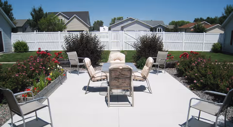 Outdoor patio area with a concrete floor, surrounded by garden beds with flowers and bushes. There are six cushioned chairs arranged around a glass-top table in the center, with additional chairs placed around the patio. A white wooden fence and residential houses are visible in the background under a clear blue sky.