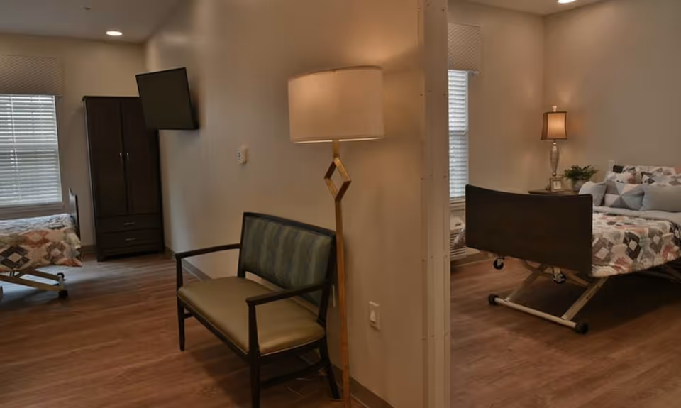Shared nursing facility bedroom with two hospital-style beds, seating, lamps, and wood flooring.