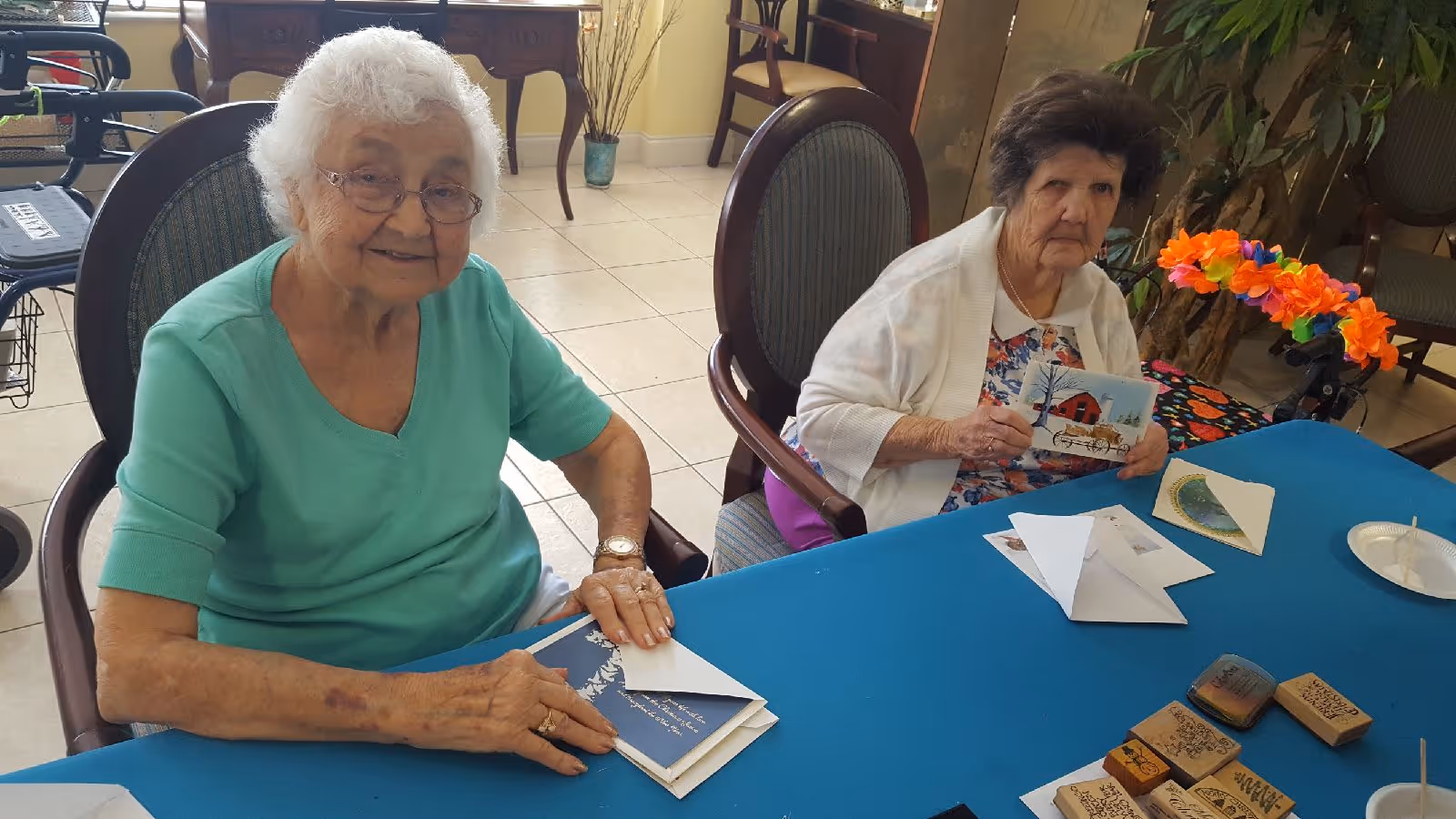 Two elderly women sitting at a table covered with a blue tablecloth, engaged in a card-making activity. One woman is smiling and wearing a green top, while the other woman is holding a card and wearing a white cardigan over a floral dress. Various crafting materials, including stamps and envelopes, are spread out on the table.