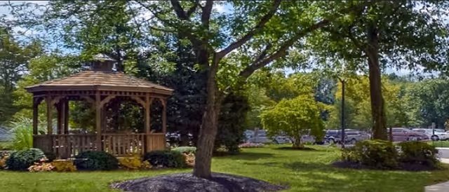A wooden gazebo surrounded by green grass, trees, and shrubs in a landscaped outdoor area with a parking lot visible in the background.