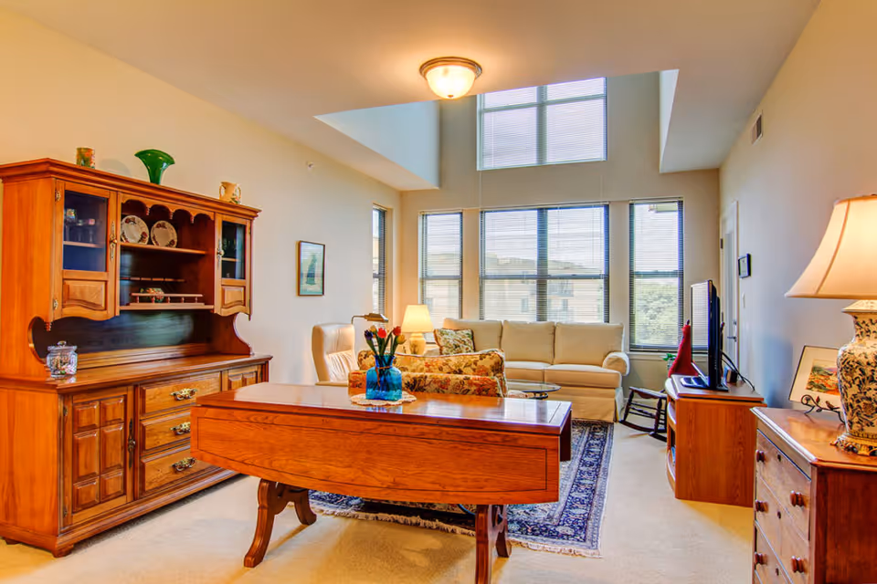 A bright living room with large windows and high ceilings. The room features a beige sofa, a floral armchair, a wooden coffee table with a blue vase of flowers, a wooden hutch with decorative plates, a TV on a wooden stand, and a lamp on a wooden dresser.