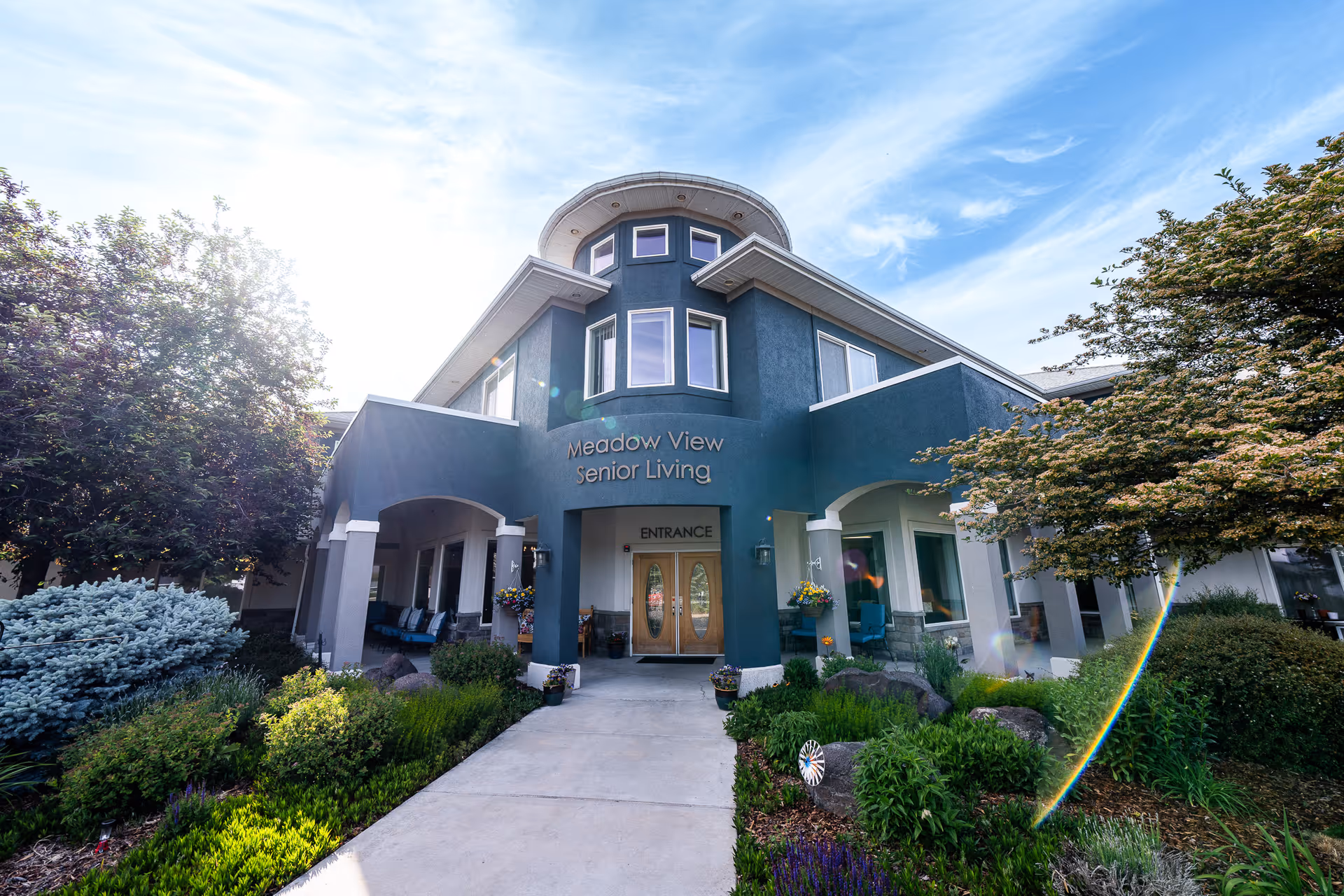 Front exterior view of Meadow View Senior Living building with a pathway leading to double wooden entrance doors, surrounded by landscaped greenery and trees under a partly cloudy blue sky.