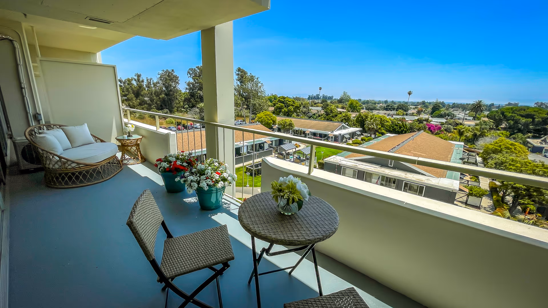 Balcony area with a small round table and two chairs, a cushioned wicker loveseat, and potted flowers. The balcony overlooks a residential area with trees and buildings under a clear blue sky.