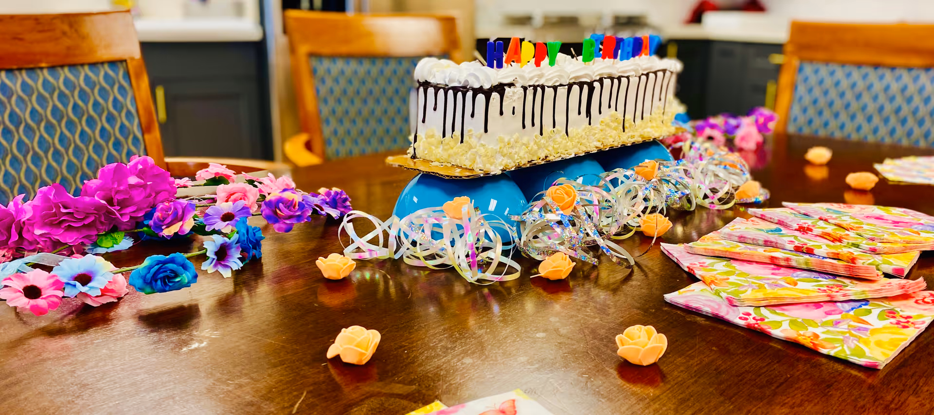 Long birthday cake on a decorated dining table with colorful flowers, ribbons, and napkins, with chairs in the background.