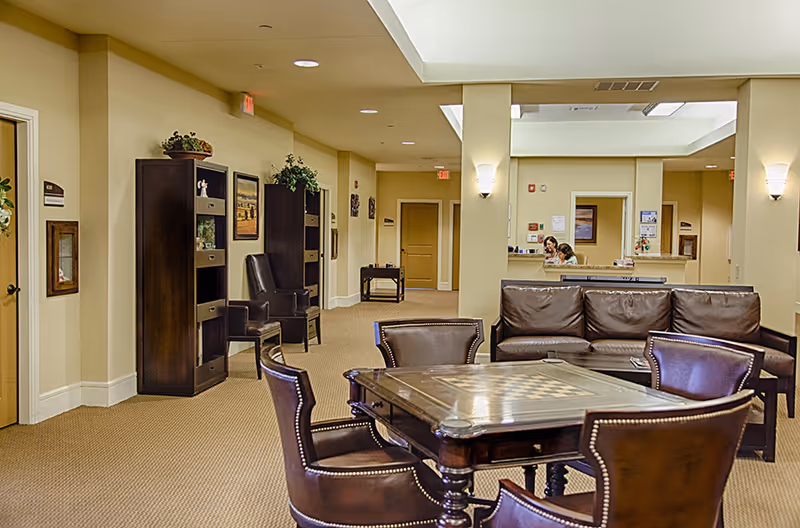 A spacious assisted living common area with a chess table surrounded by four leather chairs in the foreground. Behind the table is a brown leather couch. The room has beige walls and carpet, with dark wood shelving units and armchairs along the left wall. Two people are visible in the background near a reception desk.