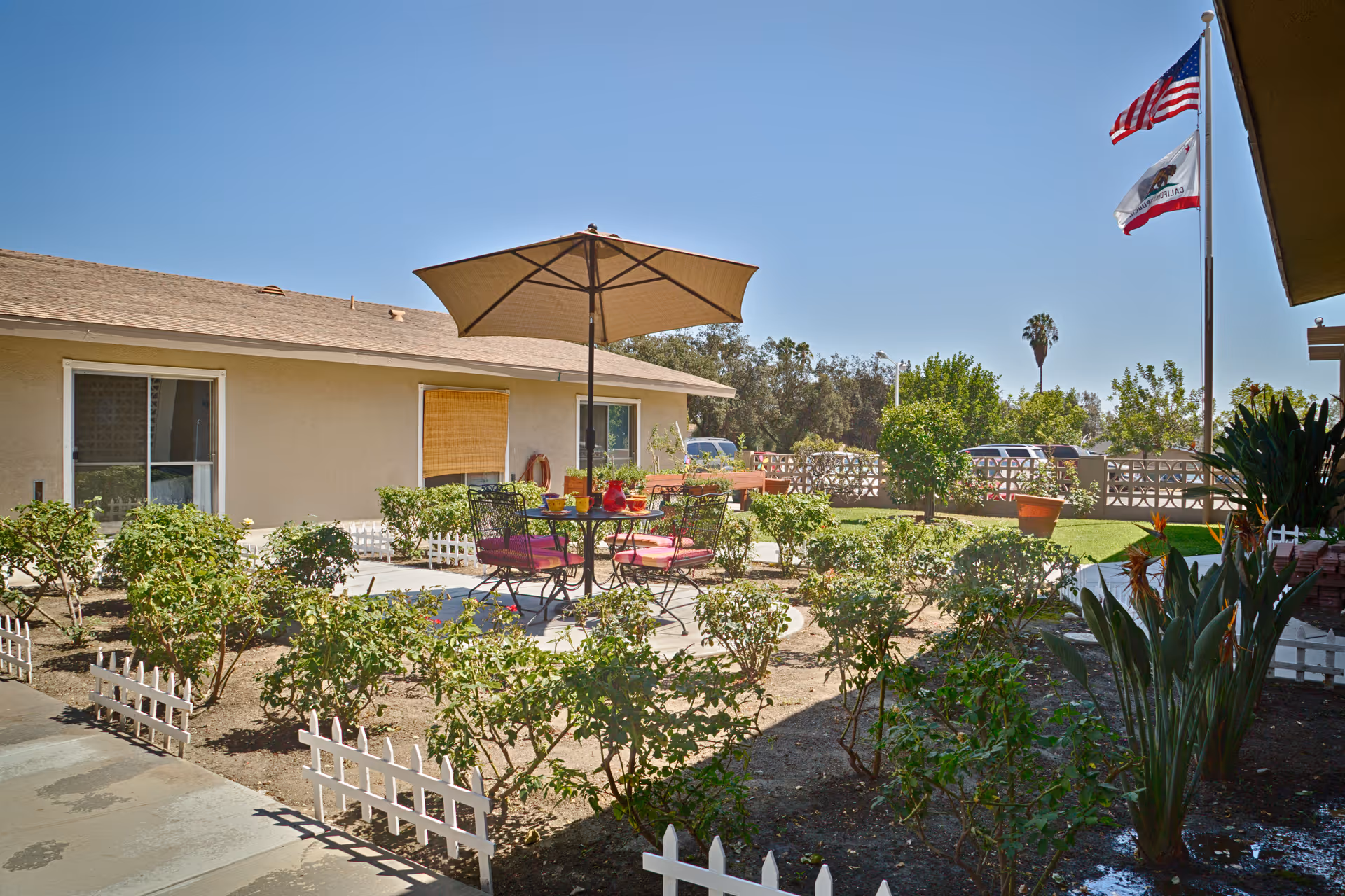 Outdoor patio area at Community Extended Care of Montclair with a round table and four chairs under a large beige umbrella. The patio is surrounded by small bushes and plants with a small white picket fence. In the background, there are two flags on a flagpole, a beige building with windows, and a clear blue sky.