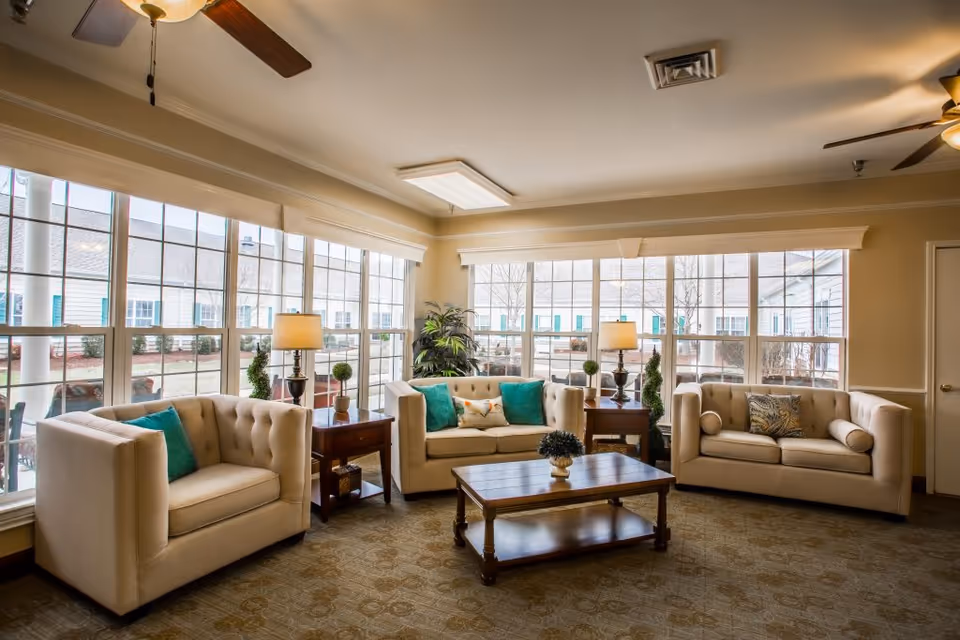 A bright and cozy living room area with three beige tufted sofas arranged around a wooden coffee table. The room has large windows letting in natural light, two side tables with lamps, and decorative plants. The ceiling features two ceiling fans with lights.