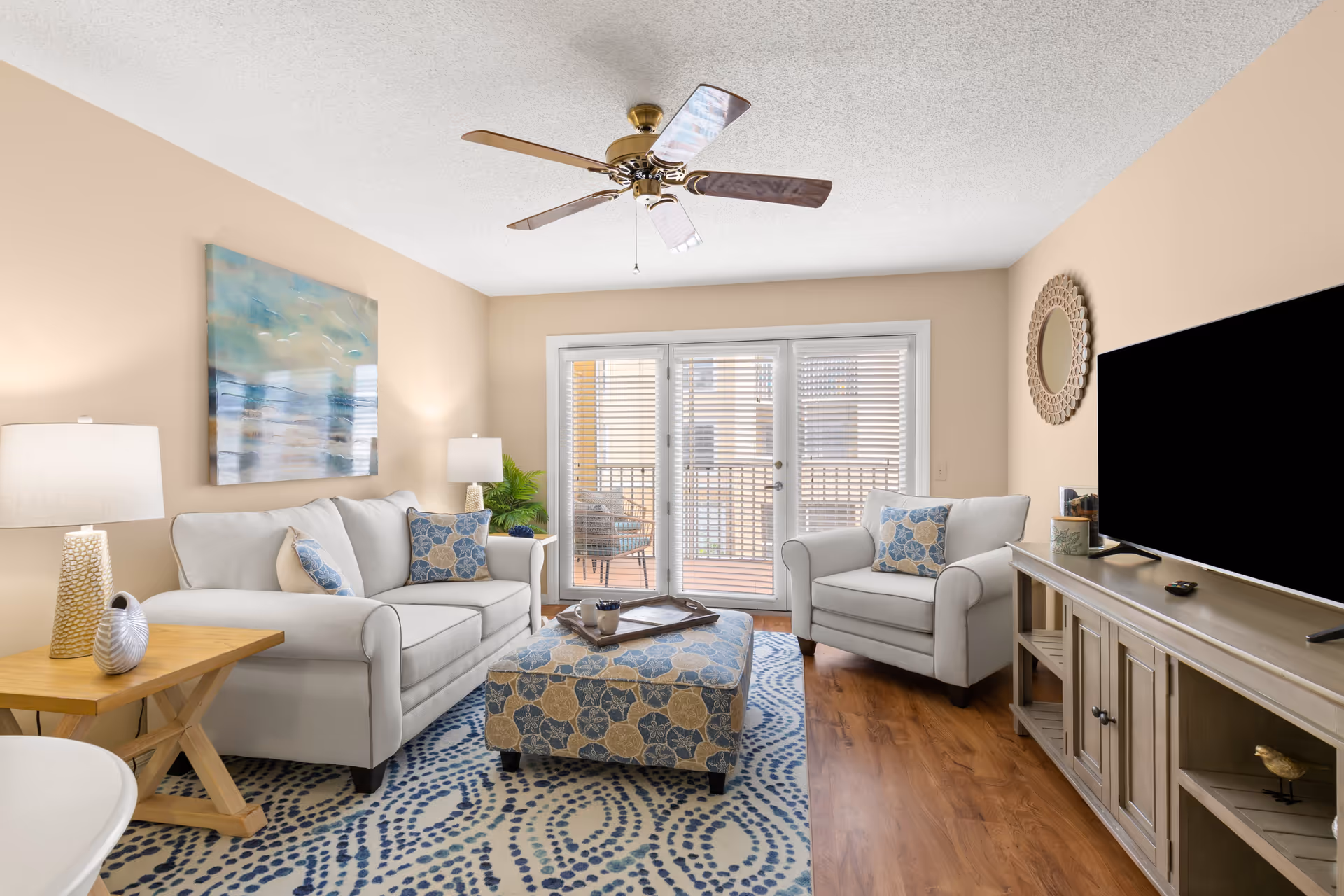 A bright living room with beige walls and wooden flooring, featuring a white sofa and armchair with blue and beige floral cushions, a matching ottoman, a wooden side table with decorative items, a ceiling fan, a large flat-screen TV on a wooden console, and glass doors leading to a balcony with outdoor seating.