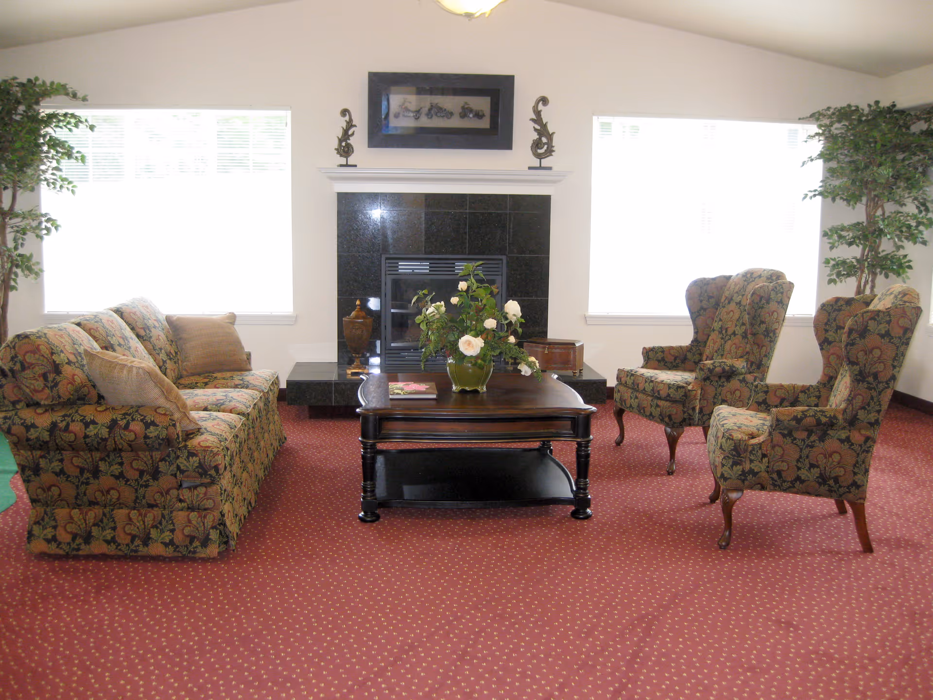 A cozy living room with a patterned sofa and two matching armchairs arranged around a dark wooden coffee table with a flower vase on it. The room has a red carpet with small yellow dots, two large windows with white blinds, and a black tiled fireplace with decorative items on the mantel.