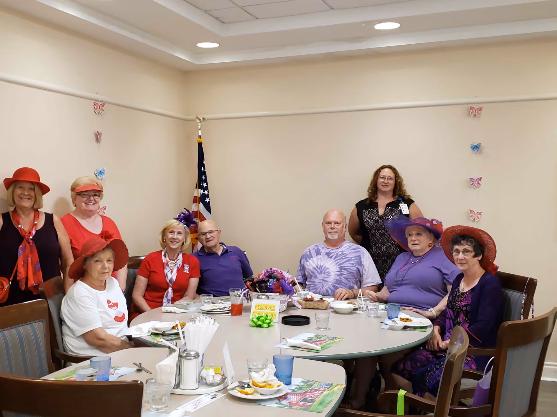 A group of eight elderly people and one staff member gathered around a large round dining table in a room decorated with butterfly wall hangings and an American flag. Some of the elderly individuals are wearing colorful hats and smiling, with plates and glasses on the table indicating a meal or event.