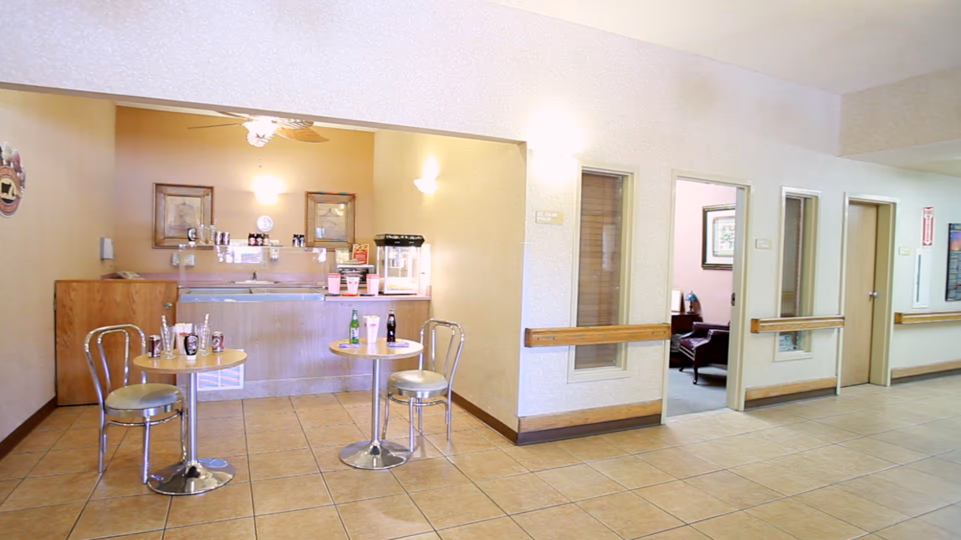 Interior view of a small dining or snack area in a senior living facility with two round tables and metal chairs. On the tables are various condiments and drink bottles. In the background, there is a counter with a popcorn machine, coffee maker, and other items. The area is well-lit with wall-mounted lights and ceiling fan. To the right, there is a hallway with doors and windows leading to other rooms.
