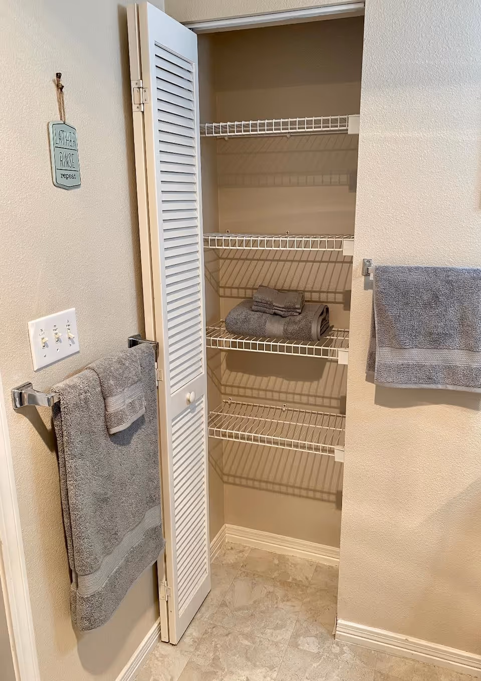 A bathroom linen closet with white wire shelves, some holding folded gray towels. The closet door is open, revealing the shelves inside. On the adjacent wall, two gray towels hang on towel racks. A small decorative sign that reads 'Lather Rinse Repeat' is hung on the wall above the towel rack. The floor is tiled in a light color.