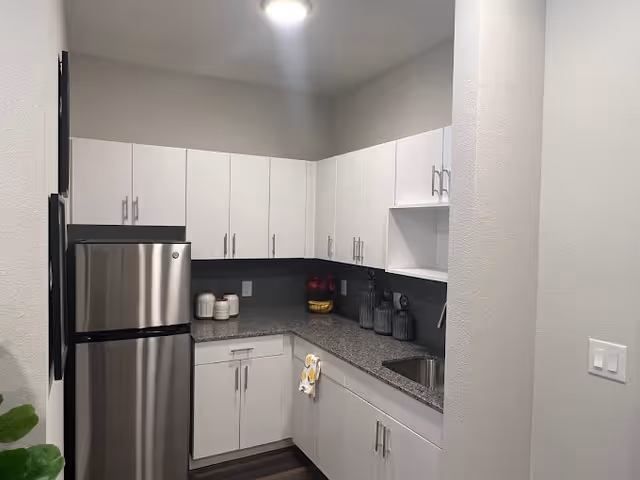 A modern kitchen with white cabinets, a stainless steel refrigerator, a granite countertop, a small sink, and decorative jars and bowls on the counter. The kitchen has a clean and minimalist design with overhead lighting.