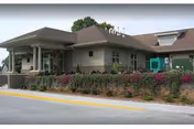 Low-rise beige building with a covered entrance, landscaped flower beds, and a driveway curb in front.