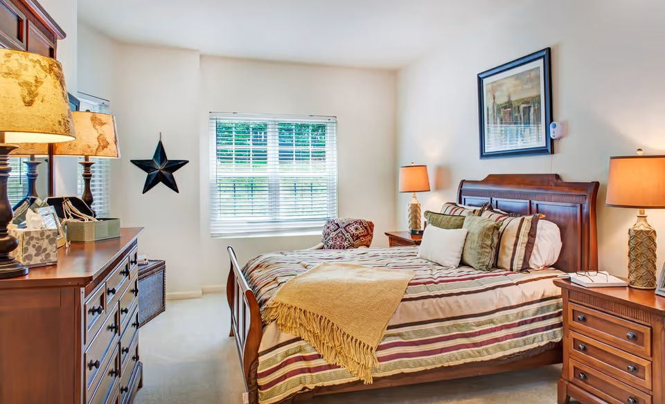 A cozy bedroom with a wooden bed frame and a striped bedspread with multiple pillows. There are two wooden nightstands on either side of the bed, each with a lamp. A framed picture hangs above the bed. A wooden dresser with a mirror and two lamps is on the left side of the room. A window with white blinds lets in natural light, and a decorative star hangs on the wall near the window.