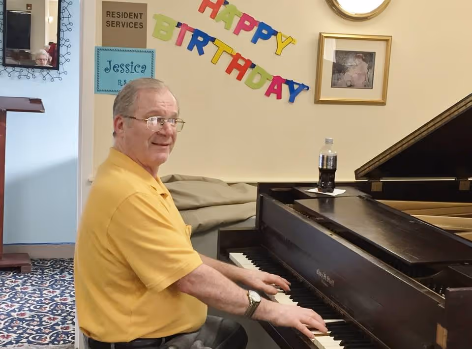 An elderly man wearing a yellow shirt and glasses is sitting at a piano, playing it and smiling. Behind him on the wall is a colorful 'Happy Birthday' banner, a framed picture, and a sign that reads 'Resident Services'. There is a bottle of soda on the piano and a patterned carpet on the floor.