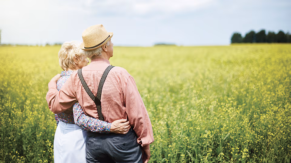 An elderly couple standing in a field of yellow flowers, embracing each other and looking into the distance under a partly cloudy sky.